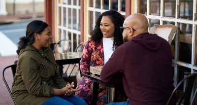 three people sitting at a table outside a cafe, smiling and chatting